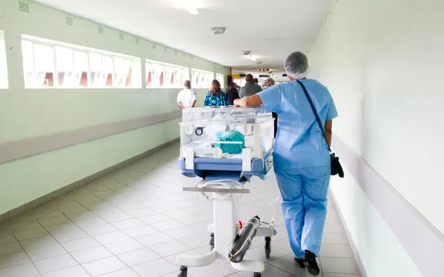 Pielęgniarka w niebieskim stroju pcha inkubator dla noworodka szpitalnym korytarzem